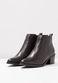 Botas de cuero negro hasta el tobillo con punta afilada, tacón bajo en bloque y cremallera lateral, que presentan una textura suave y un diseño minimalista.