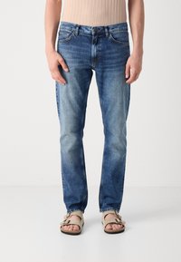 Jeans negre închis cu un design cu picioare drepte, având o ușoară uzură și decolorare. Accompanied de sandale de culoare deschisă.
