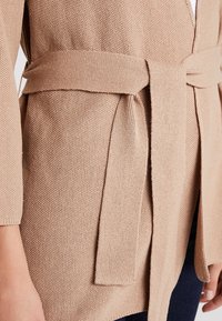 Ljusbeige stickad cardigan med en strukturerad yta, öppen framdel och matchande tygbälte. Har långa ärmar och en avslappnad passform.