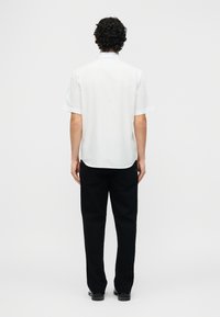 Άνδρας με σγουρά μαλλιά που στέκεται στραμμένος προς τα πίσω, φοράει λευκό πουκάμισο με κοντά μανίκια και μαύρο παντελόνι, σε απλό ανοιχτό φόντο.