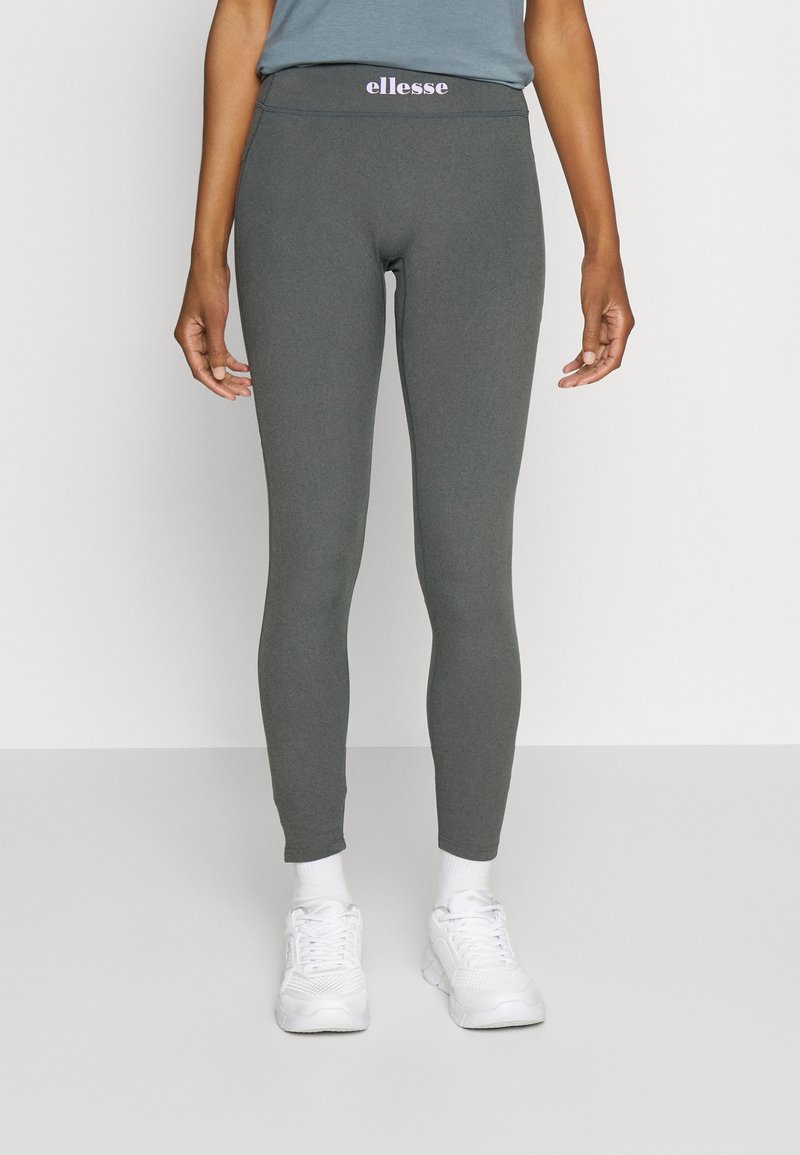 Legging gris avec un design ajusté, fabriqué en tissu extensible, présentant un logo violet visible sur la ceinture et associé à des baskets blanches.