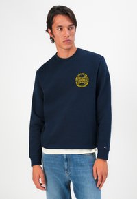 Mladic nosi tamnoplavi duksericu sa žutim Tommy Jeans logom na grudima, svijetloplave traperice i bijelu majicu ispod na bijeloj pozadini.