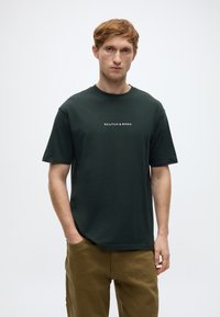 Σκούρο πράσινο βαμβακερό μπλουζάκι με στρογγυλή λαιμόκοψη και κοντά μανίκια, με λευκό κείμενο λογότυπου τοποθετημένο στο κέντρο του στήθους.