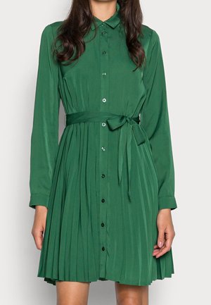 Kvinde iført en langærmet grøn plisseret midi-kjole med knapper foran og et bundet bælte i taljen.