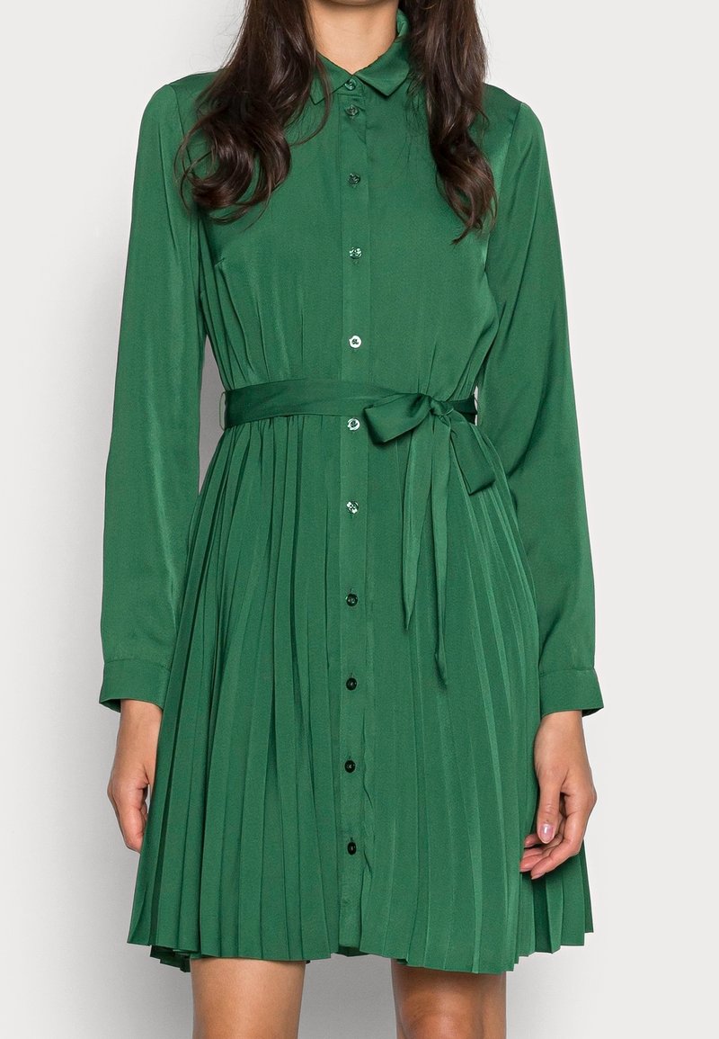 Femme portant une robe midi plissée verte à manches longues avec des boutons sur le devant et une ceinture nouée à la taille.