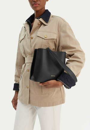 Person trägt eine beige Jacke mit schwarzem Kragen und Manschetten, hält eine schwarze Lederhandtasche mit Schulterriemen und goldenen Akzenten.