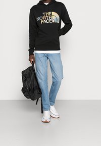 Felpa nera con il logo "The North Face", jeans azzurri chiari, sneaker bianche e uno zaino nero, presentati su uno sfondo neutro.