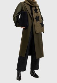 Abrigo largo verde oliva con mangas negras, combinado con una bufanda marrón con estrellas negras, bolso negro, pantalones negros de pierna ancha y botines negros.