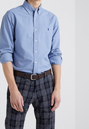 Homme portant une chemise bleu clair boutonnée avec les manches retroussées, une ceinture marron et un pantalon à carreaux foncé sur un fond uni.