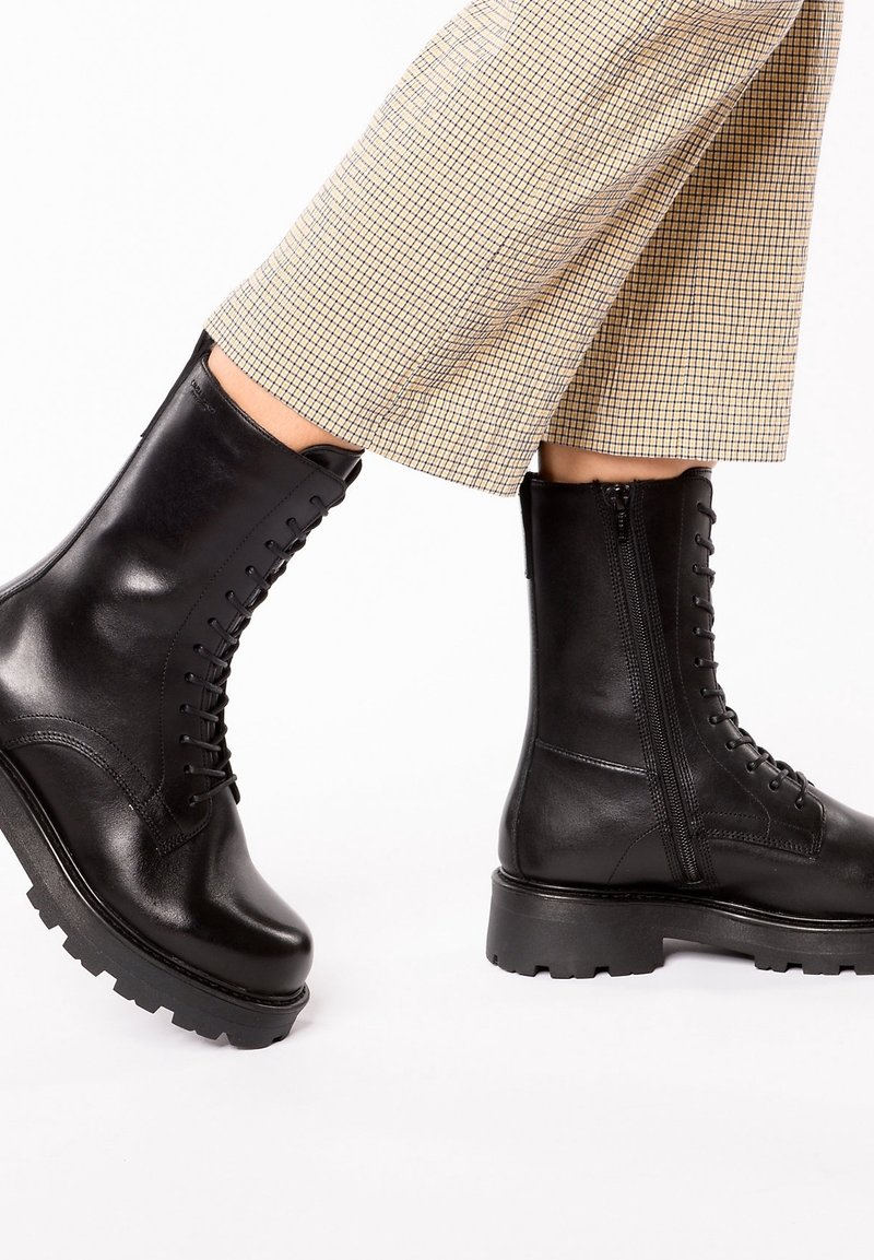 Bottes en cuir noir à lacets avec fermeture éclair latérale portées avec un pantalon court à carreaux beige sur un fond clair.