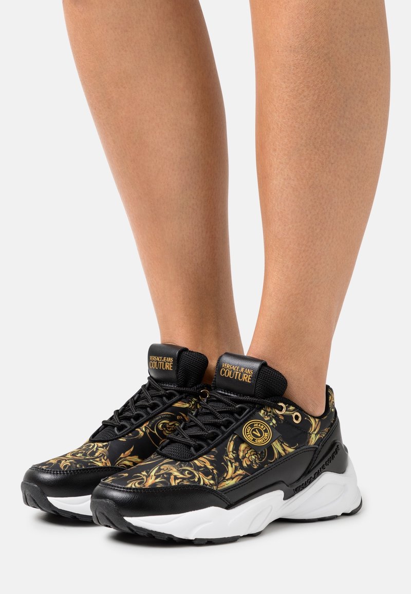 Svarta sneakers med guldblommönster, strukturerat tyg, vit sula och svarta snören, med logodetaljer på tungan och sidan.