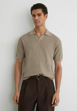 Mees, kes kannab beežikat tekstuuriga lühikeste varrukatega polosärki, mis on pistetud tumesinistesse pükstesse, millel on hõbedane pandlaga vöö, seistes valge seina taustal.