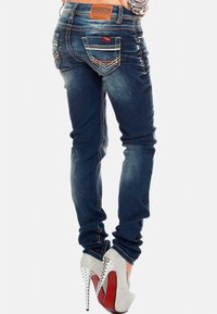 Jeans in denim blu snelli con lavaggio scuro, caratterizzati da tasche posteriori con cuciture decorative e una patch in pelle marrone. Indossati con tacchi alti argentati.