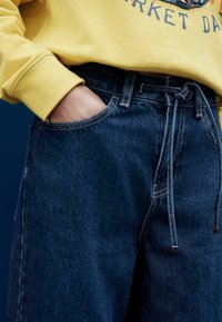 Pantalones vaqueros de color azul oscuro con cintura con cordón, combinados con una sudadera amarilla. Incluyen bolsillos delanteros y costuras en contraste.