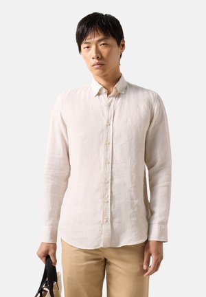 Junger Mann, der ein beigefarbenes Hemd mit langen Ärmeln und Knöpfen sowie khakifarbene Hosen trägt, hält eine schwarz-beige Tragetasche vor einem schlichten Hintergrund.