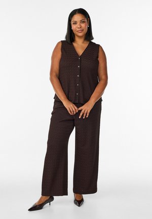 Woman standing wearing sleeveless brown button-front top, matching wide-leg pants, and black pointed-toe heels against white background.