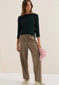 Donkergroene longsleeve top, beige elastische taillebroek en lichtroze corduroy pet. Soepele texturen, casual pasvorm en relaxte stijl.