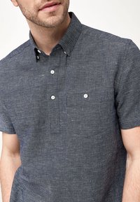 Chemise à manches courtes, boutonnée, en tissu bleu foncé à tissage texturé. Comprend une poche poitrine et des boutons blancs.