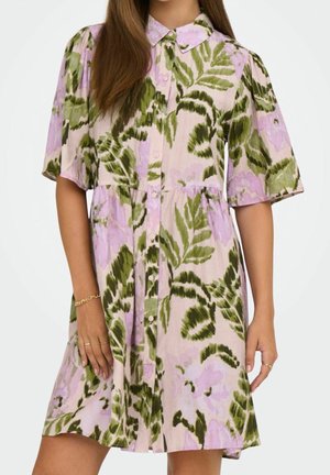 Woman wearing a button-up dress with green and purple floral patterns, short sleeves, and a collar, standing against a plain background.