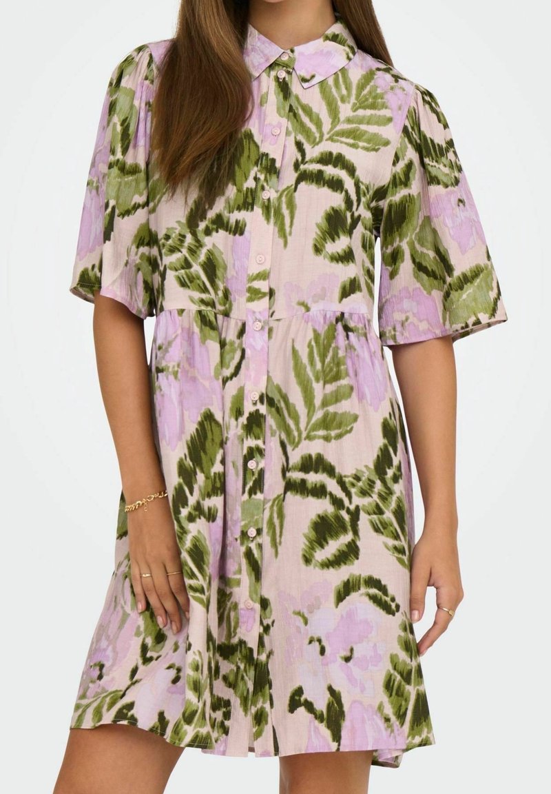 Femme portant une robe boutonnée avec des motifs floraux verts et violets, des manches courtes et un col, debout devant un fond uni.