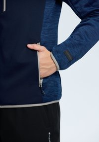 Giacca softshell blu navy con pannello laterale testurizzato blu, tasca con zip e dettagli grigi. Tessuto liscio con un design aderente per un uso attivo.