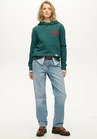 Groene hoodie met witte trekkoorden, met een rood logo. Gecombineerd met lichtblauwe jeans en bruine veterschoenen. Gestreept shirt eronder.