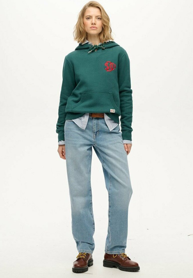 Lyshåret kvinde iført en teal hoodie, stribet skjorte, lyseblå jeans, brun bælte og brune snørebåndsstøvler, der står foran en hvid baggrund.