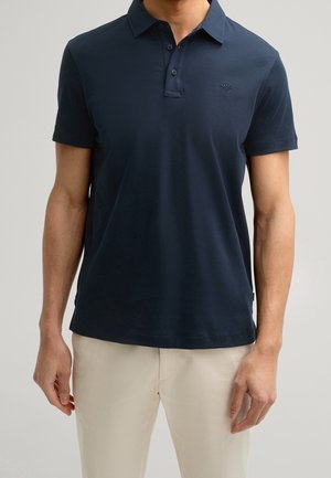 Mann trägt dunkelblaues kurzärmeliges Poloshirt mit kleinem gesticktem Logo, kombiniert mit beigen Hosen, steht vor neutralem Hintergrund.