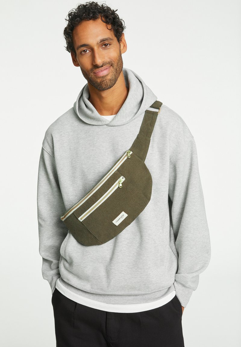 Banane vert olive avec deux poches zippées, en tissu texturé et un label de logo blanc. Portée en travers du corps avec une sangle ajustable.