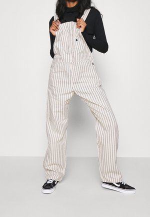 Beige gestreepte overalls met verstelbare bandjes, een voorzak en wijde pijpen, gecombineerd met zwarte sneakers met witte zolen.