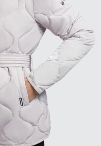 Lys grå quilted jakke med bælte i taljen, der har en glat overflade og fremtrædende bølgemønstre. Inkluderer en sidelomme og logodetalje.