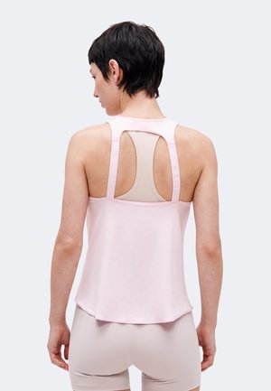 Žena s krátkymi čiernymi vlasmi, ktorá má na sebe svetloružové tielko bez rukávov cez béžovú športovú podprsenku, doplnené o béžové šortky, zobrazená zozadu.