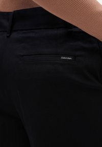 Črne bombažne hlače z zadnjim žepom, na katerem je prišita nalepka z napisom "Calvin Klein." Pas je raven in diskreten.