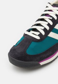 Tealfärgade och marinblå mockaskor med vita ränder, texturerad yta och en lila accent på sulan. Snörningen är krämfärgad med en klassisk design.