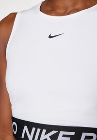 Biały sportowy biustonosz z okrągłym dekoltem, z czarnym logo Nike umieszczonym centralnie oraz szerokim czarnym pasem z białym napisem na dole.