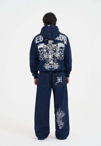 Dunkelblaues Denim-Hoodie und weite Hosen. Das Hoodie hat ein großes grafisches Design mit Totenköpfen und Rosen; die Hosen sind mit kunstvoller Drachengestickung versehen.