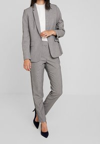 Grijs getailleerd colbert met bijpassende, op maat gemaakte broek. De stof is glad; de broek heeft accentueringen aan de zijkant. Draagt bij een wit shirt en marineblauwe hakken.