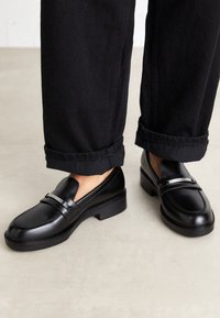 Mocassins en cuir noir à finition lisse, avec une large bride argentée proéminente et un talon bas et épais. Portés avec un pantalon noir retroussé.