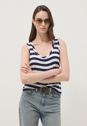 Femme aux longs cheveux bruns portant un haut sans manches rayé, un jean bleu clair, une ceinture marron, des boucles d’oreilles créoles en or et des lunettes de soleil rectangulaires, les bras croisés.