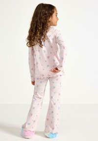 Pigiama rosa con motivo floreale, maniche lunghe e pantaloni svasati; tessuto morbido con bordi arricciati; indossato con pantofole pelose multicolori.