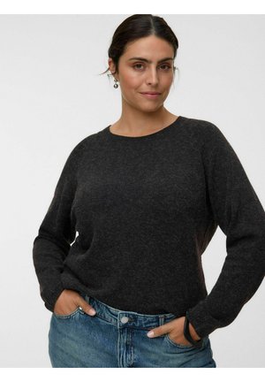 Femme aux cheveux foncés, portant un pull gris foncé et un jean bleu, debout les mains dans les poches devant un fond clair et uni.