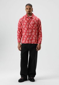 Rood overhemd met knopen en een patroon met witte motieven, voorzien van een borstzak, gecombineerd met zwarte broek met wijde pijpen en zwarte schoenen.