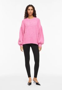 Pull rose tricoté à manches larges, avec un motif floral texturé, accompagné d'un pantalon noir ajusté et de talons noirs à bout pointu.