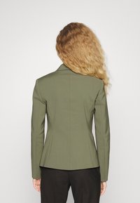 Blazer aderente verde oliva con silhouette strutturata, rever affilati e tessuto liscio; presenta un retro sagomato senza chiusure visibili.