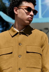 Chaqueta marrón con textura y cuello, dos bolsillos frontales y cierre de botones, combinada con gafas de sol ámbar contra un fondo colorido.