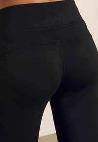 Leggings sportivi neri con una texture liscia, vita alta e dettagli di cucitura in vita. Presentano un logo discreto vicino alla parte posteriore.
