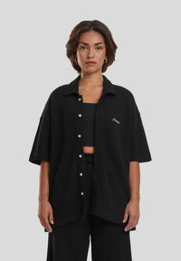 Svart ribbet knapper-skjorte med krage, kortermet, og hvit brodering på venstre side, kombinert med en svart crop top og matchende bukser.