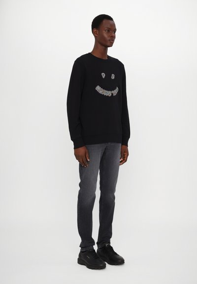 Sudadera negra con un diseño de cara sonriente, con texto multicolor. Combinada con jeans oscuros y zapatillas negras. Textura suave.