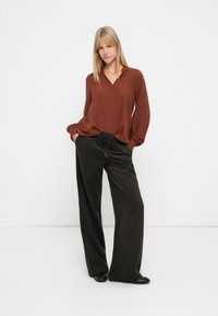 Bruine blouse met lange mouwen en een V-hals, gecombineerd met hoge taille, wijde donkere broeken. Gladde stoffen en een ontspannen pasvorm.