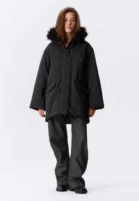 Parka neagră cu glugă căptușită cu blană sintetică, închidere frontală cu butoane și două buzunare laterale. Pantaloni largi din material texturat în două tonuri, cu un model subtil.